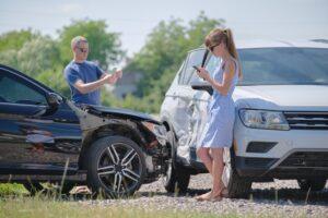 Two people standing next to their cars after a car accident. The woman is contacting her insurance company.